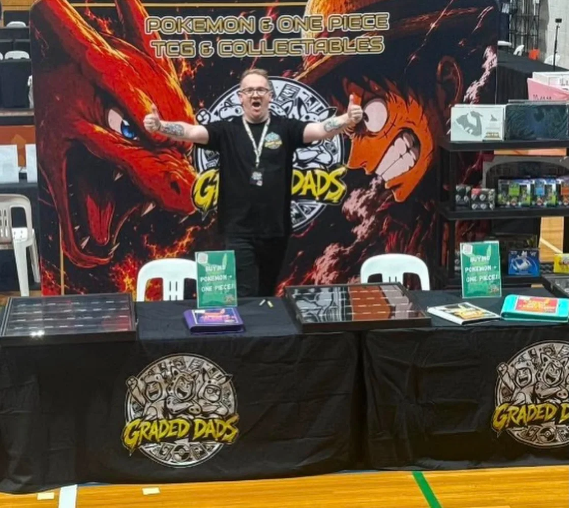 A team member in a Graded Dads shirt gives two thumbs up behind the stall, in front of the Charizard and Luffy banner
