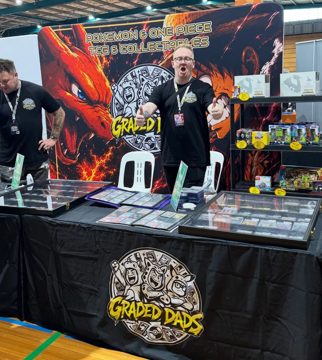 Two team members behind the Graded Dads stall, with display cases of graded cards, binders and shelves of sealed product